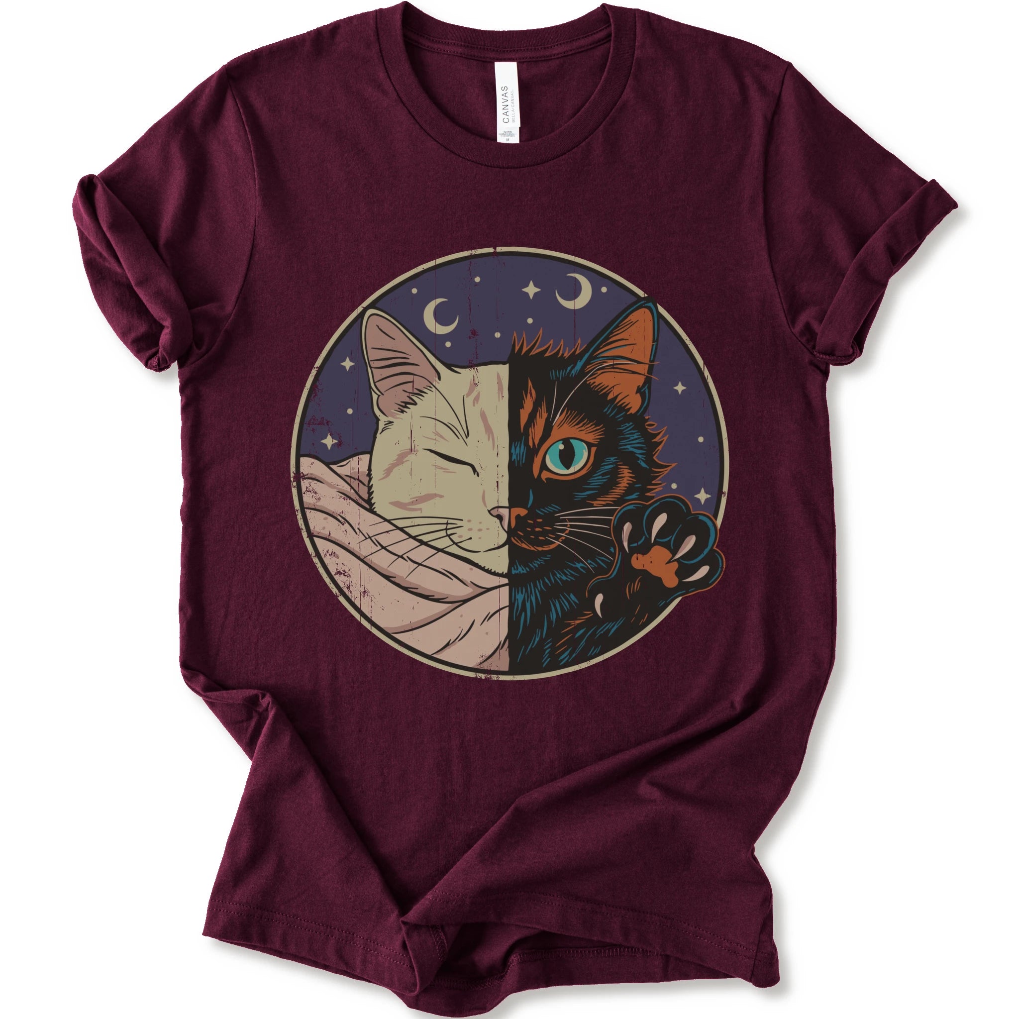 A maroon short-sleeve crewneck t-shirt featuring a circular graphic print of a split cat design, showing one half as a sleeping cream-colored cat wrapped in a blanket and the other half as a dark black cat with a glowing blue eye and paw resting on its head.