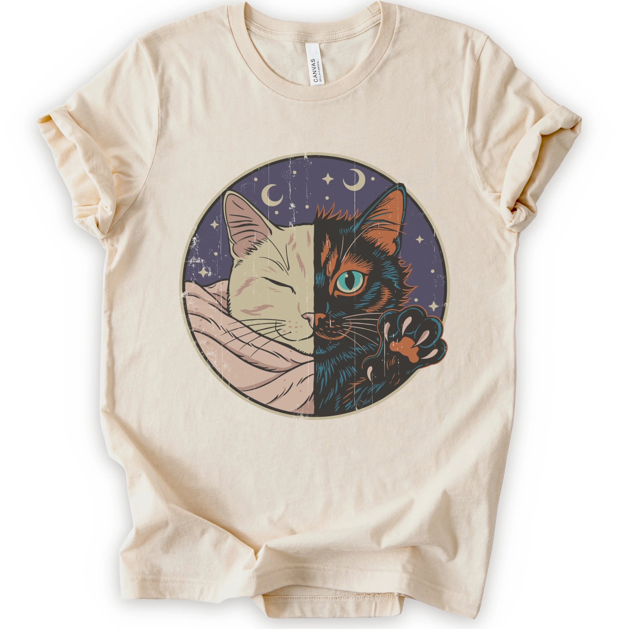 A natural short-sleeve crewneck t-shirt featuring a circular graphic print of a split cat design, showing one half as a sleeping cream-colored cat wrapped in a blanket and the other half as a dark black cat with a glowing blue eye and paw resting on its head.