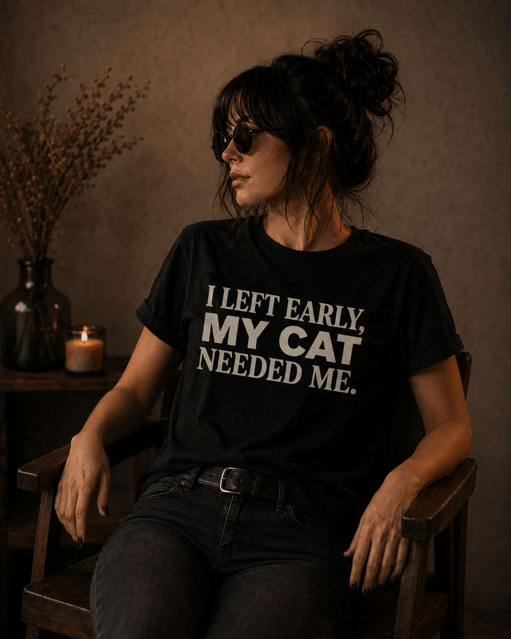 A woman wearing a black graphic t-shirt with the text 'I LEFT EARLY, MY CAT NEEDED ME.' in white, sitting in a rustic wooden chair in a dimly lit room.