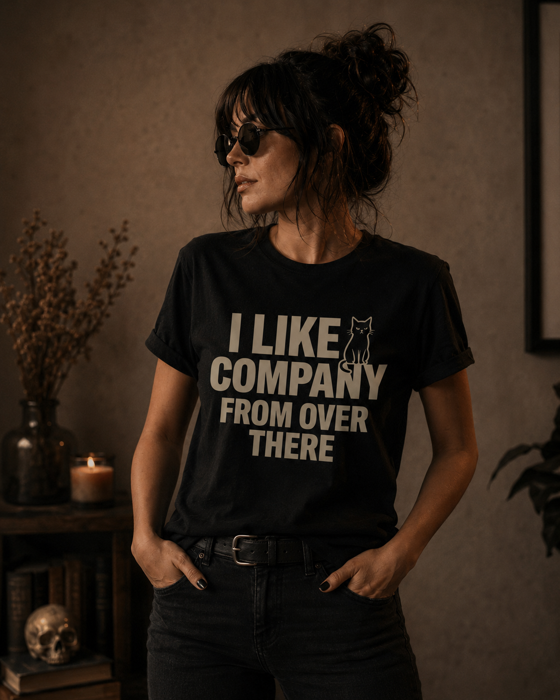 A woman wearing a black graphic t-shirt with the text 'I LIKE COMPANY FROM OVER THERE' and a small cat silhouette, styled in a moody, warm-toned indoor setting.