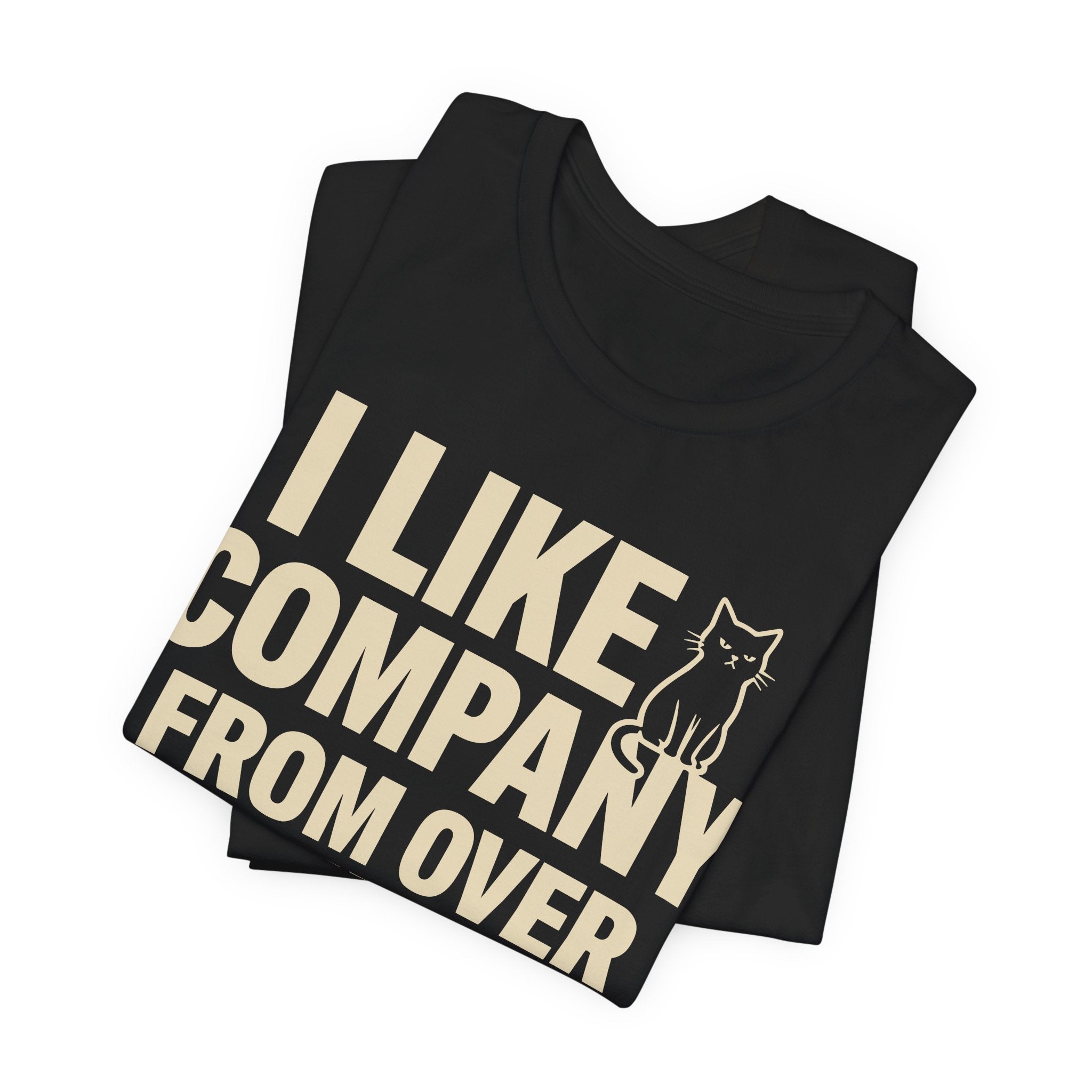 A folded black short-sleeve t-shirt with a beige graphic print that reads "I Like Company From Over There" with an unimpressed sitting cat