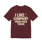 A maroon short-sleeve t-shirt with a beige graphic print that reads "I Like Company From Over There" with an unimpressed sitting cat