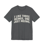 A asphalt short-sleeve crewneck t-shirt featuring a graphic with Distressed cream text reading “I LIKE THREE BEINGS, ONE JUST MEOWS.” 