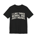 A black short-sleeve crewneck t-shirt featuring a graphic with Distressed cream text reading “I LIKE THREE BEINGS, ONE JUST MEOWS.” 