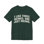 A forest short-sleeve crewneck t-shirt featuring a graphic with Distressed cream text reading “I LIKE THREE BEINGS, ONE JUST MEOWS.” 