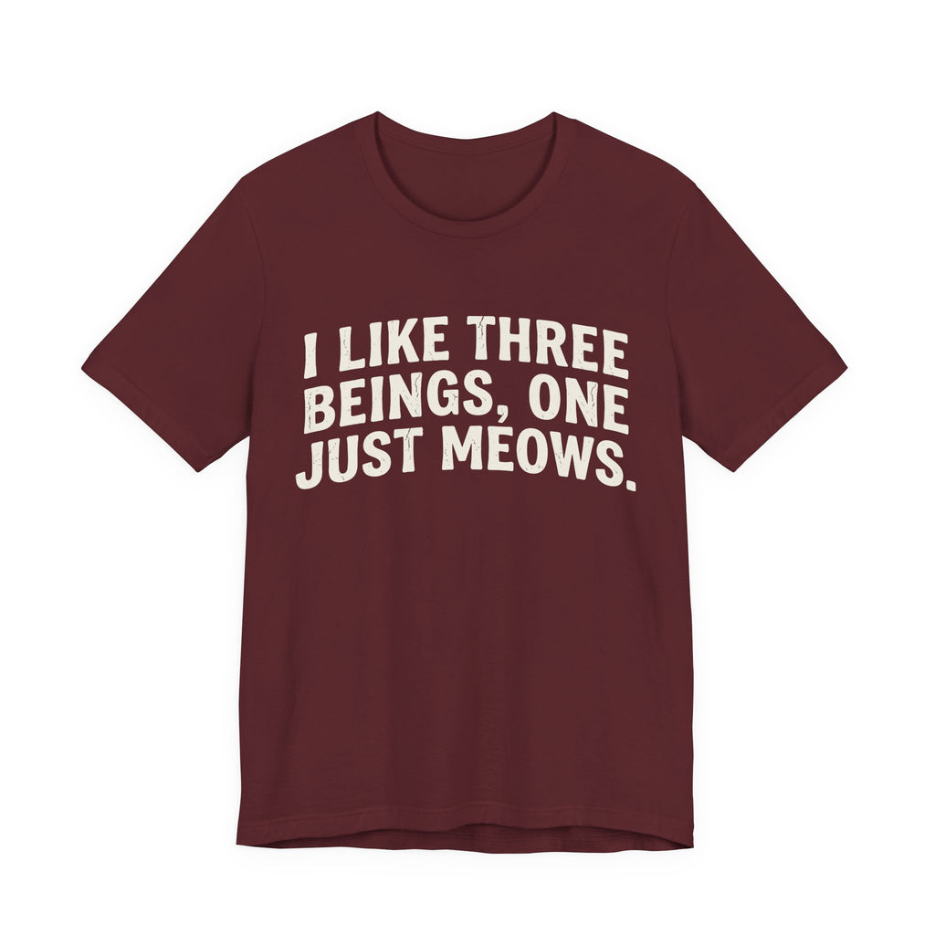 A maroon short-sleeve crewneck t-shirt featuring a graphic with Distressed cream text reading “I LIKE THREE BEINGS, ONE JUST MEOWS.” 