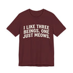 A maroon short-sleeve crewneck t-shirt featuring a graphic with Distressed cream text reading “I LIKE THREE BEINGS, ONE JUST MEOWS.” 