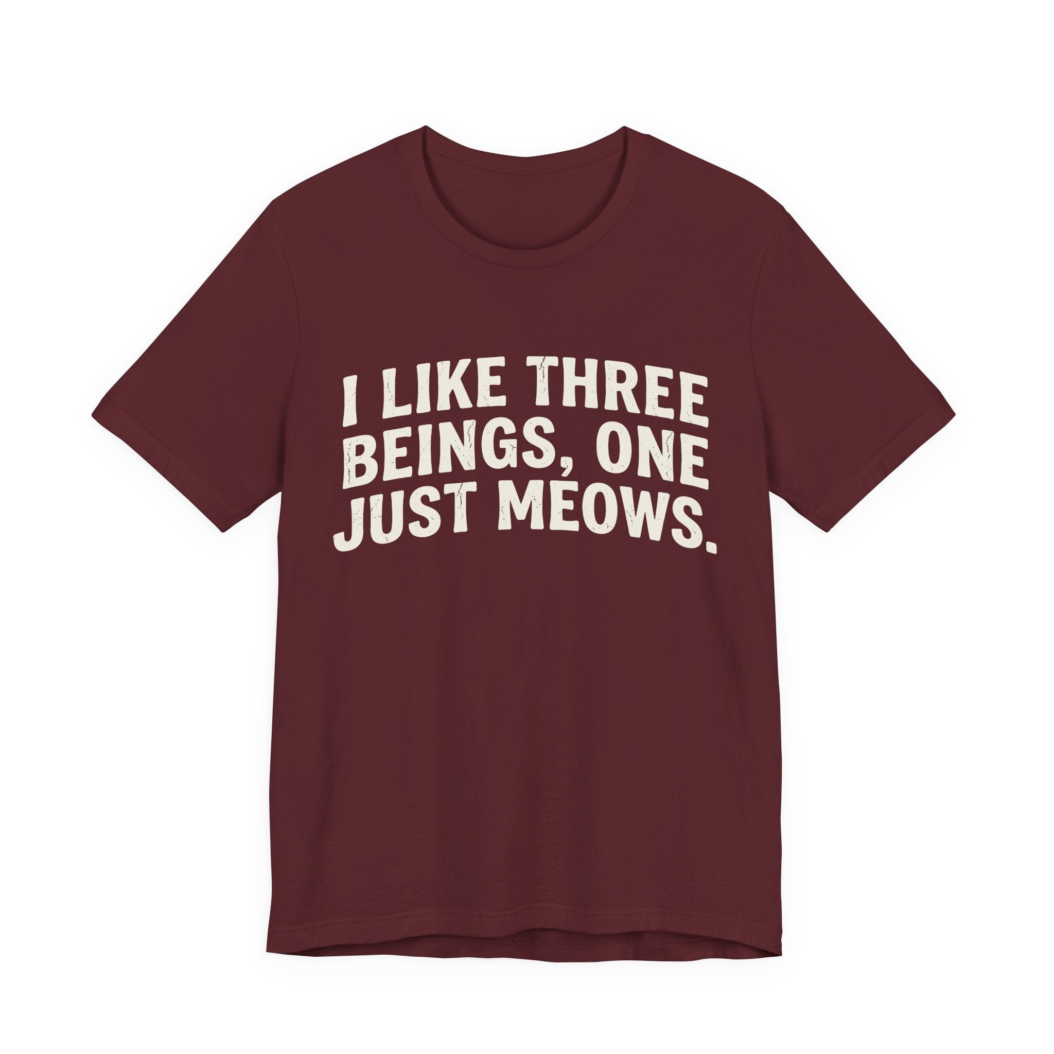A maroon short-sleeve crewneck t-shirt featuring a graphic with Distressed cream text reading “I LIKE THREE BEINGS, ONE JUST MEOWS.” 
