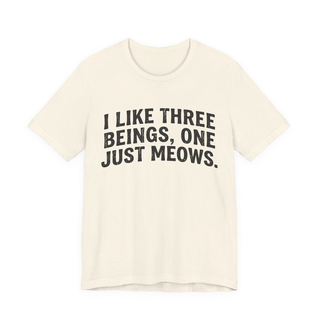 A natural short-sleeve crewneck t-shirt featuring a graphic with Distressed black text reading “I LIKE THREE BEINGS, ONE JUST MEOWS.” 