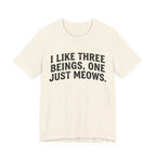 A natural short-sleeve crewneck t-shirt featuring a graphic with Distressed black text reading “I LIKE THREE BEINGS, ONE JUST MEOWS.” 