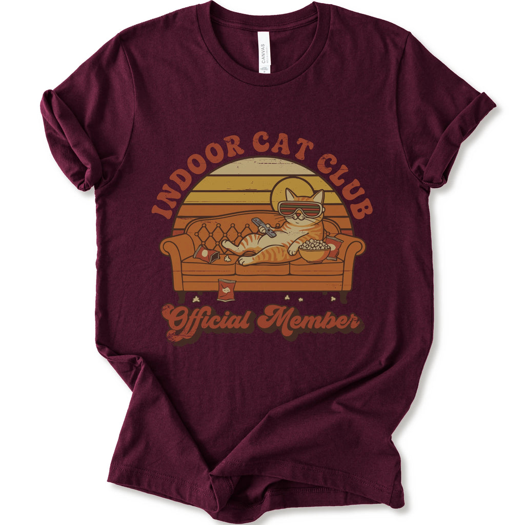 A maroon short-sleeve crewneck t-shirt featuring a retro-style graphic of a cat lounging on a couch with the text 'INDOOR CAT CLUB' and 'Official Member' in orange-brown lettering.