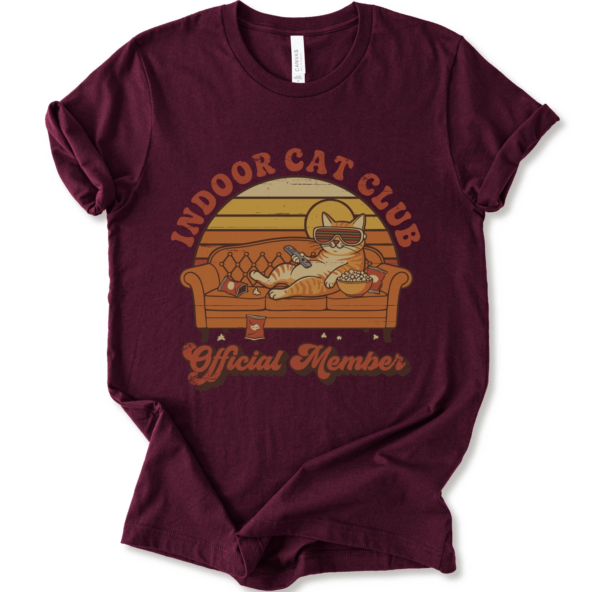 A maroon short-sleeve crewneck t-shirt featuring a retro-style graphic of a cat lounging on a couch with the text 'INDOOR CAT CLUB' and 'Official Member' in orange-brown lettering.