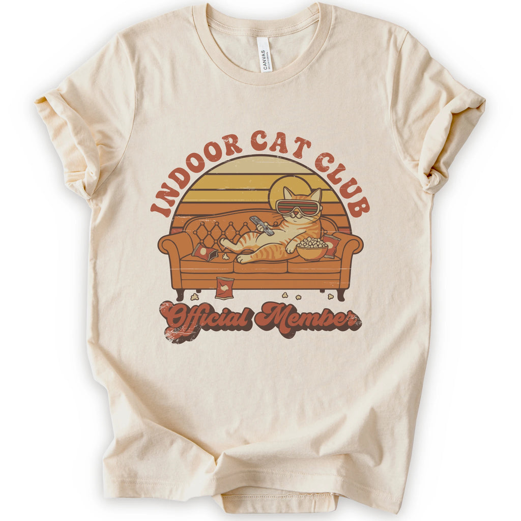 A natural short-sleeve crewneck t-shirt featuring a retro-style graphic of a cat lounging on a couch with the text 'INDOOR CAT CLUB' and 'Official Member' in orange-brown lettering.