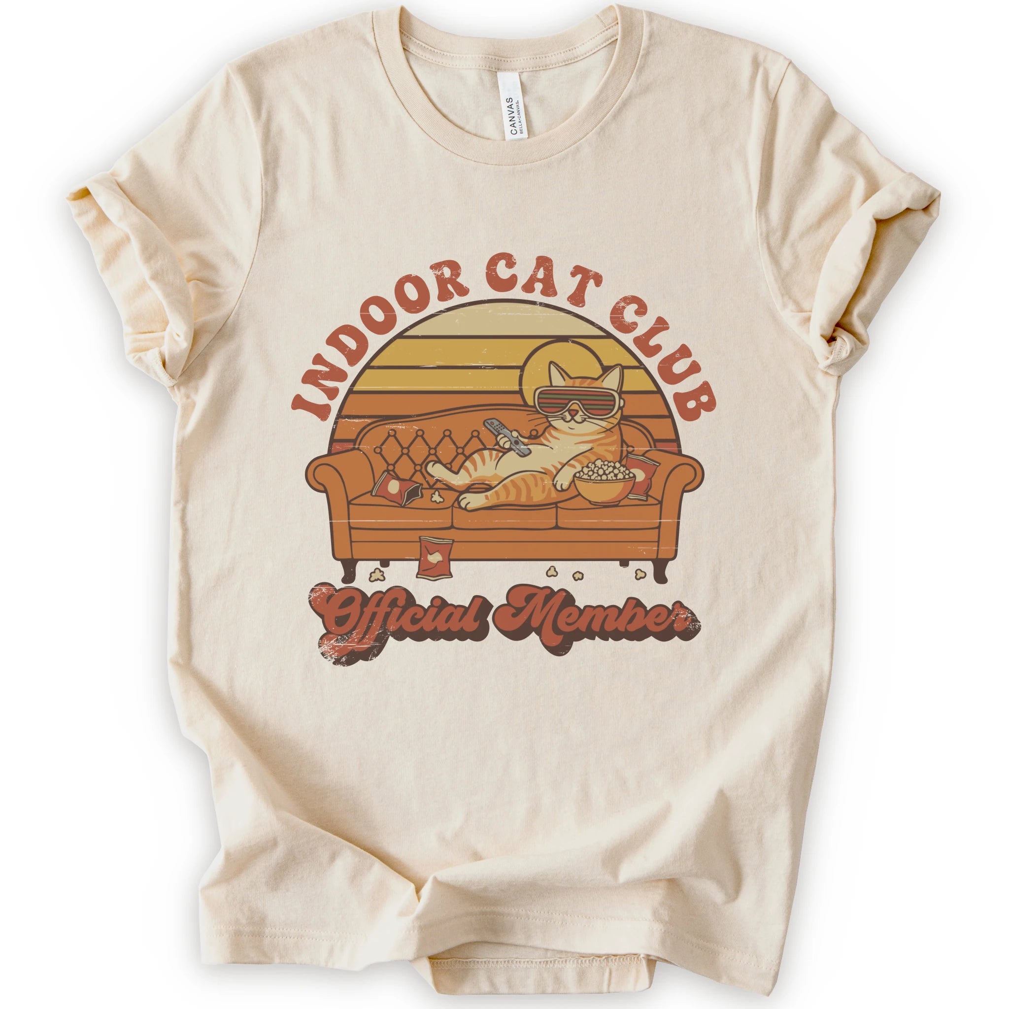 A natural short-sleeve crewneck t-shirt featuring a retro-style graphic of a cat lounging on a couch with the text 'INDOOR CAT CLUB' and 'Official Member' in orange-brown lettering.
