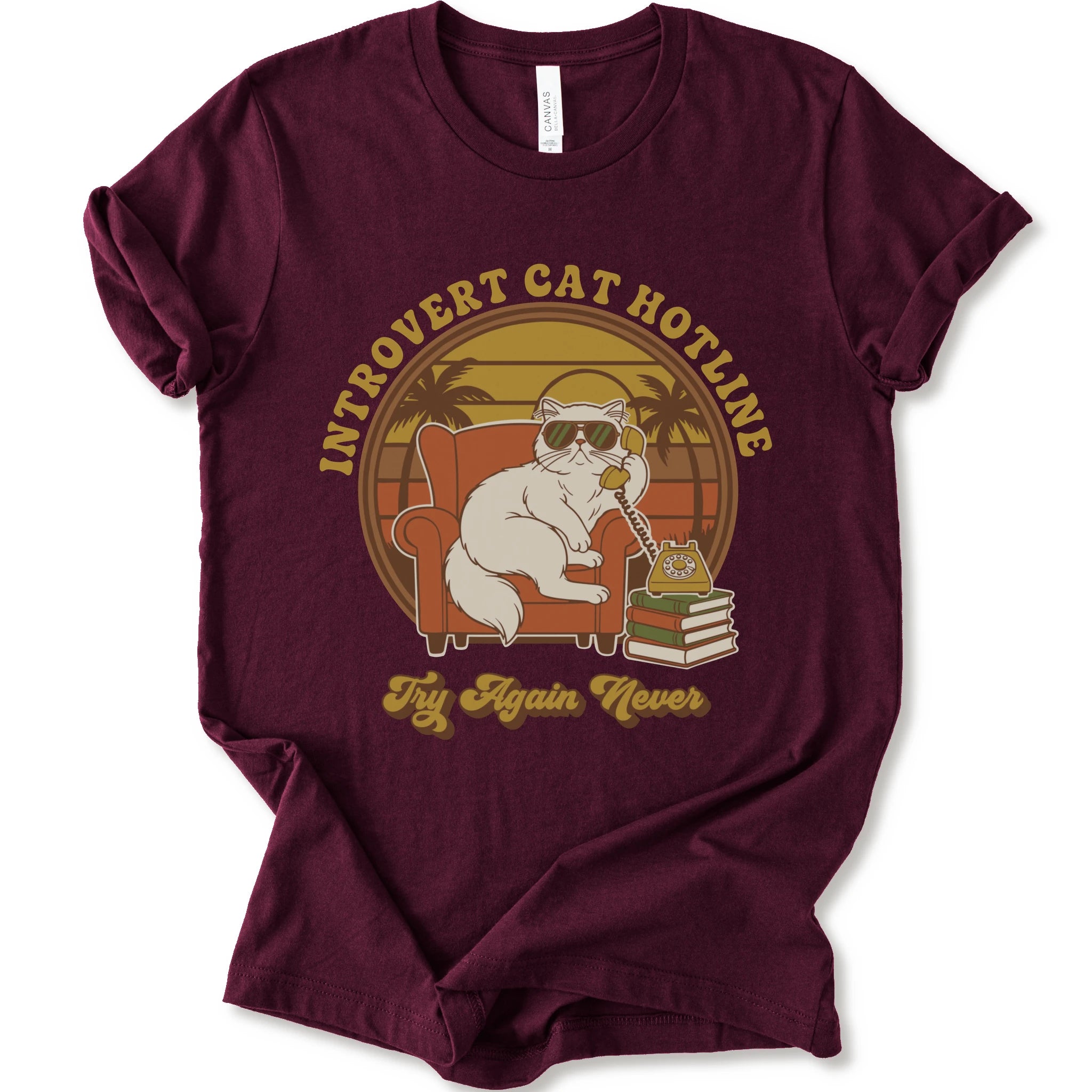 A maroon short-sleeve crewneck t-shirt with a retro-style graphic of a white cat wearing sunglasses and sitting in an orange armchair, surrounded by the text 'INTROVERT CAT HOTLINE' and 'Try Again Never'.