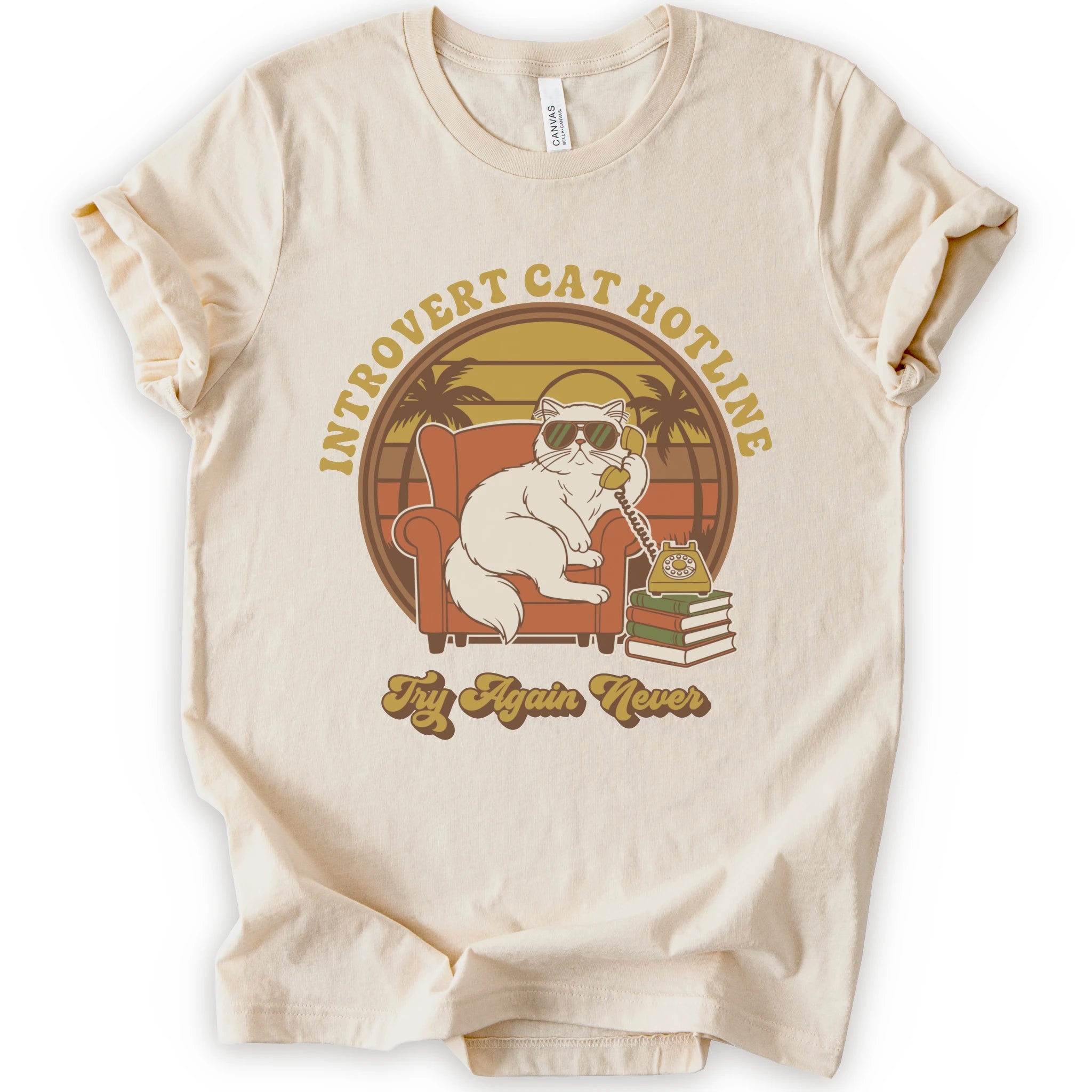A natural short-sleeve crewneck t-shirt with a retro-style graphic of a white cat wearing sunglasses and sitting in an orange armchair, surrounded by the text 'INTROVERT CAT HOTLINE' and 'Try Again Never'.