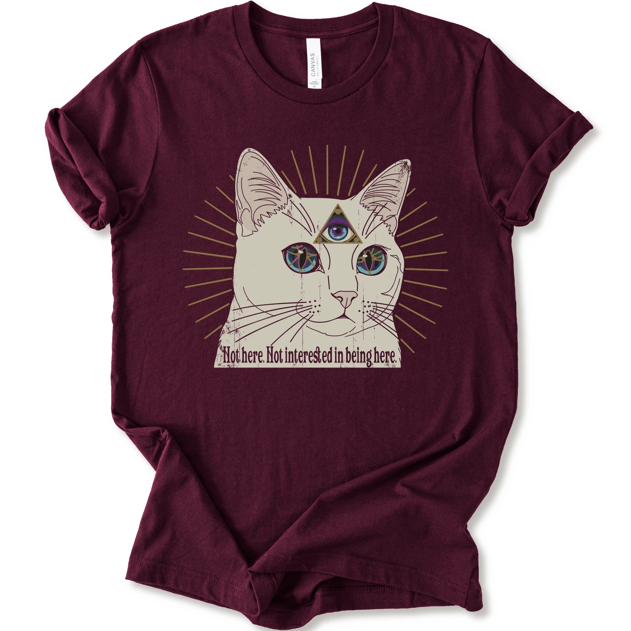 A maroon short-sleeve crewneck t-shirt featuring a graphic of a white cat with a third eye and gold sunburst rays, with the text 'Not here. Not interested in being here.