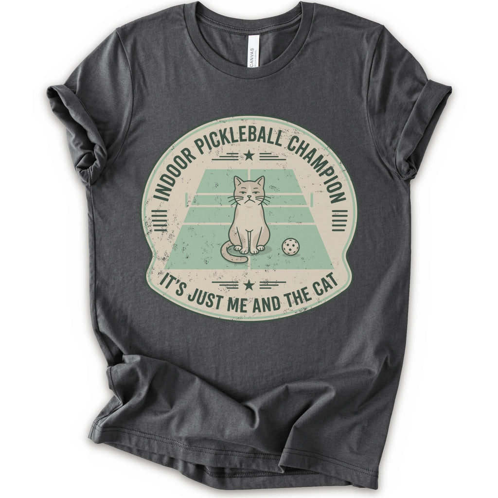 A asphalt short-sleeve crewneck t-shirt with a distressed circular graphic on the front, features a cartoon cat playing pickleball on a court, with the text 'INDOOR PICKLEBALL CHAMPION' and 'IT'S JUST ME AND THE CAT'.