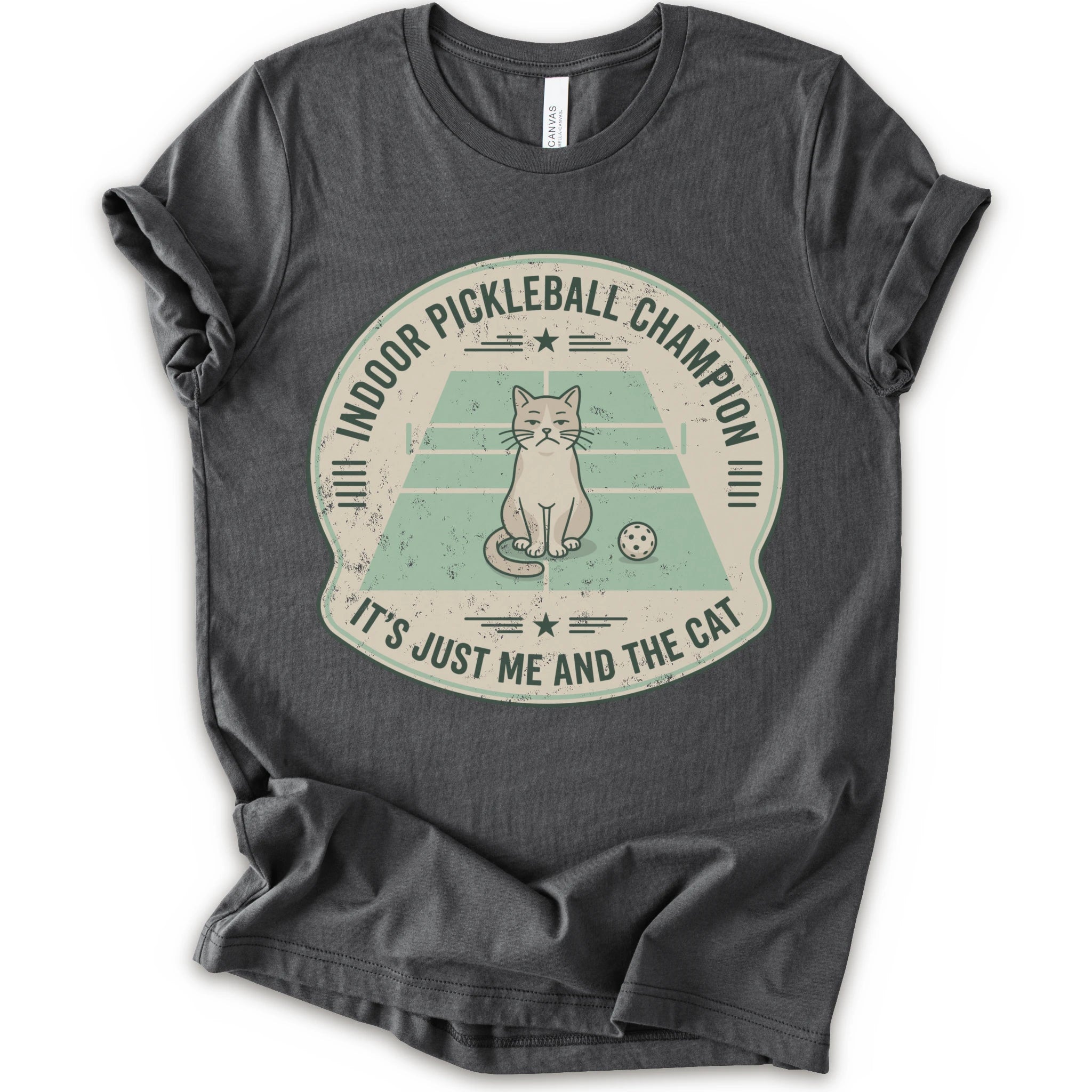 A asphalt short-sleeve crewneck t-shirt with a distressed circular graphic on the front, features a cartoon cat playing pickleball on a court, with the text 'INDOOR PICKLEBALL CHAMPION' and 'IT'S JUST ME AND THE CAT'.