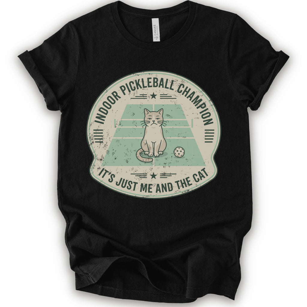 A black short-sleeve crewneck t-shirt with a distressed circular graphic on the front, features a cartoon cat playing pickleball on a court, with the text 'INDOOR PICKLEBALL CHAMPION' and 'IT'S JUST ME AND THE CAT'.