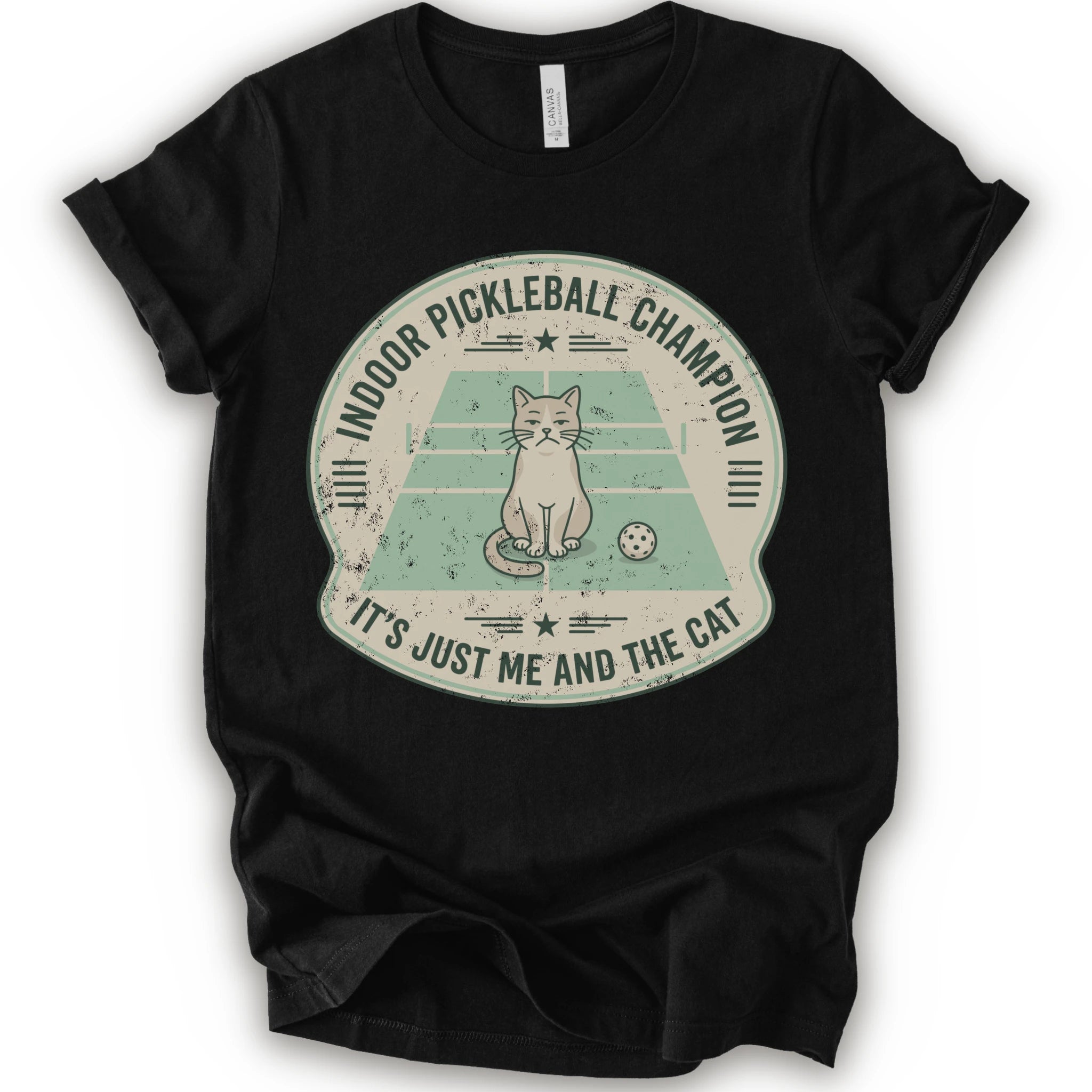 A black short-sleeve crewneck t-shirt with a distressed circular graphic on the front, features a cartoon cat playing pickleball on a court, with the text 'INDOOR PICKLEBALL CHAMPION' and 'IT'S JUST ME AND THE CAT'.