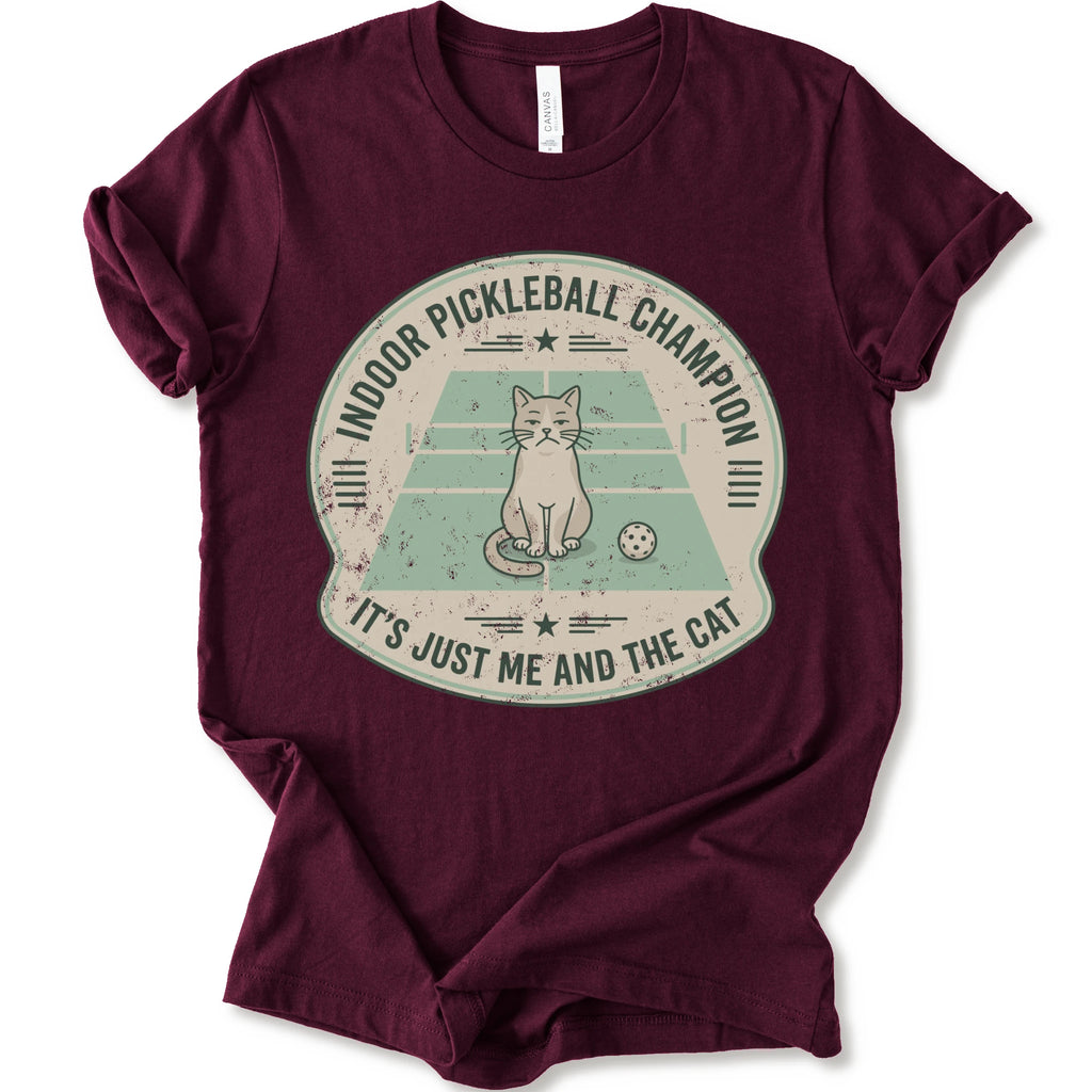 A maroon short-sleeve crewneck t-shirt with a distressed circular graphic on the front, features a cartoon cat playing pickleball on a court, with the text 'INDOOR PICKLEBALL CHAMPION' and 'IT'S JUST ME AND THE CAT'.