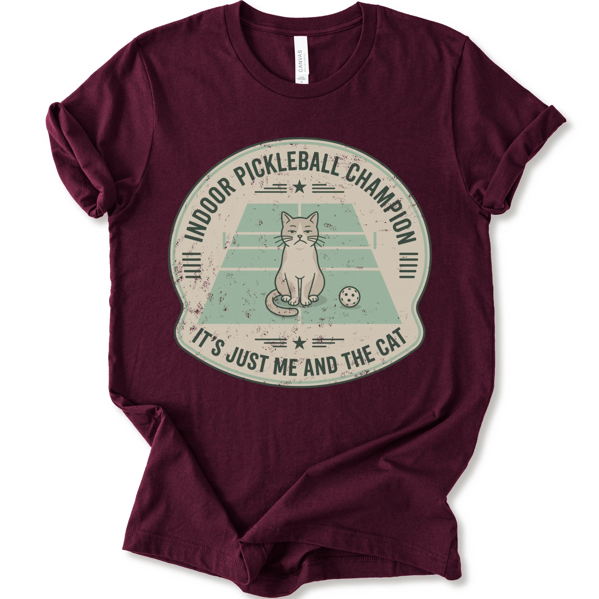 A maroon short-sleeve crewneck t-shirt with a distressed circular graphic on the front, features a cartoon cat playing pickleball on a court, with the text 'INDOOR PICKLEBALL CHAMPION' and 'IT'S JUST ME AND THE CAT'.