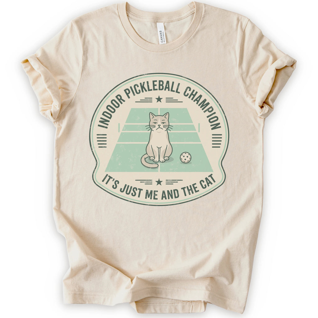 A natural short-sleeve crewneck t-shirt with a distressed circular graphic on the front, features a cartoon cat playing pickleball on a court, with the text 'INDOOR PICKLEBALL CHAMPION' and 'IT'S JUST ME AND THE CAT'.