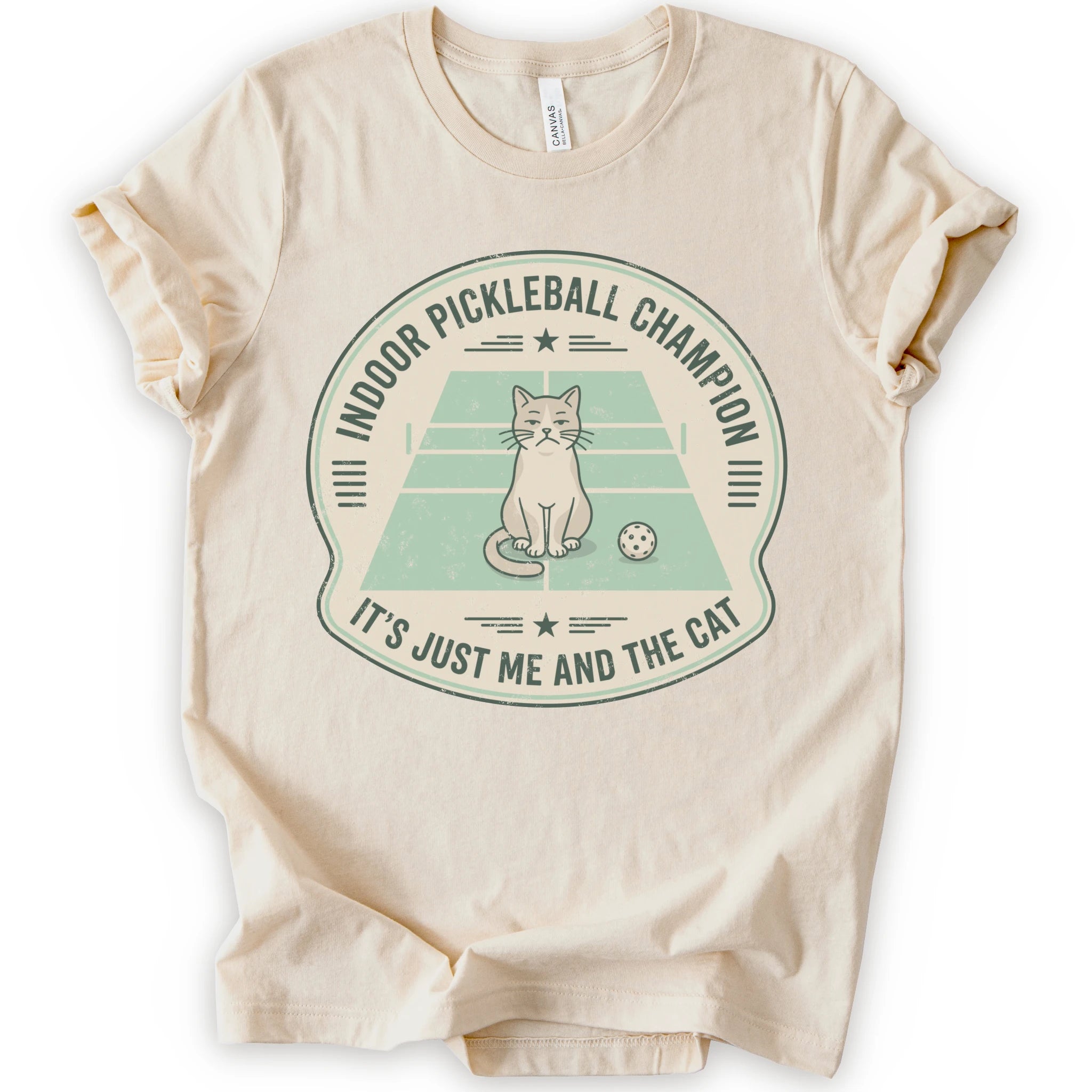 A natural short-sleeve crewneck t-shirt with a distressed circular graphic on the front, features a cartoon cat playing pickleball on a court, with the text 'INDOOR PICKLEBALL CHAMPION' and 'IT'S JUST ME AND THE CAT'.