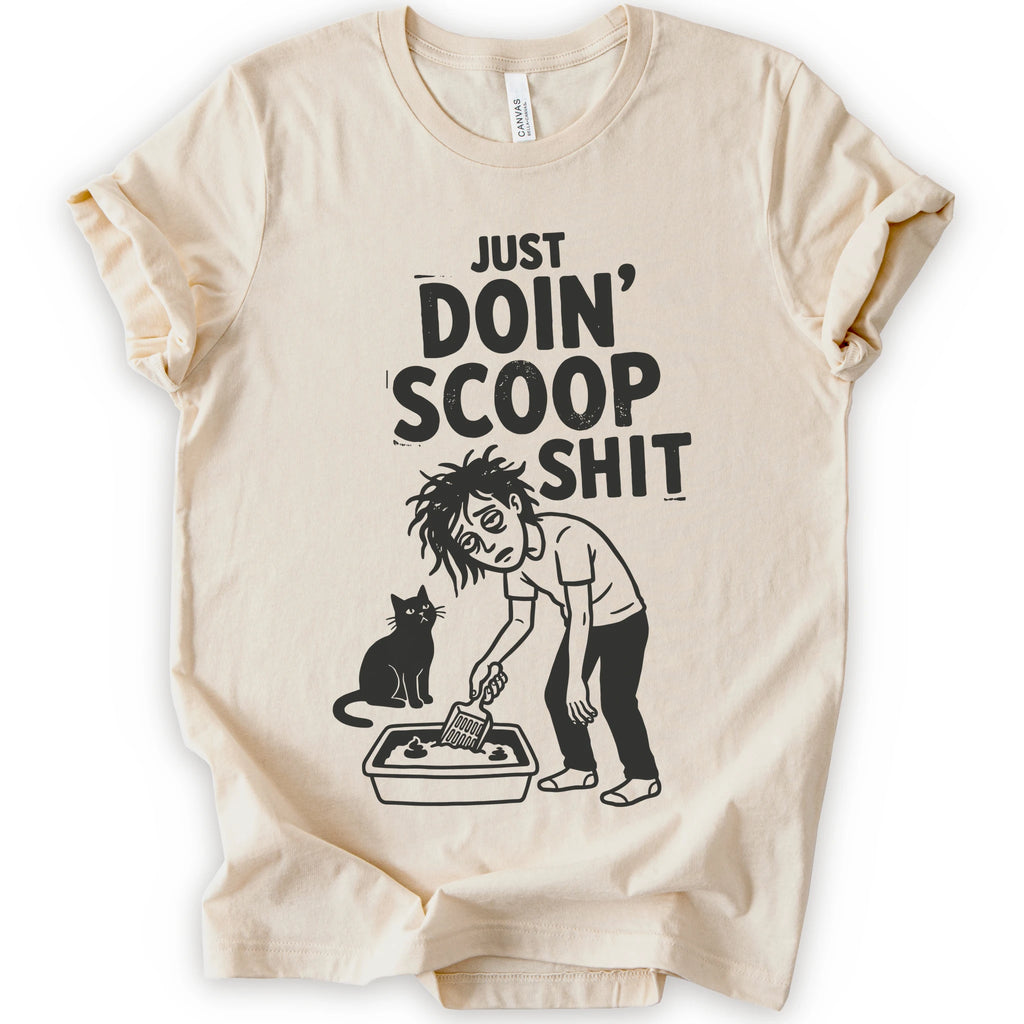 A natural short-sleeve crewneck t-shirt featuring a graphic with 
“Just Doin’ Scoop Shit” text above a tired cat owner cleaning a litter box beside a seated cat