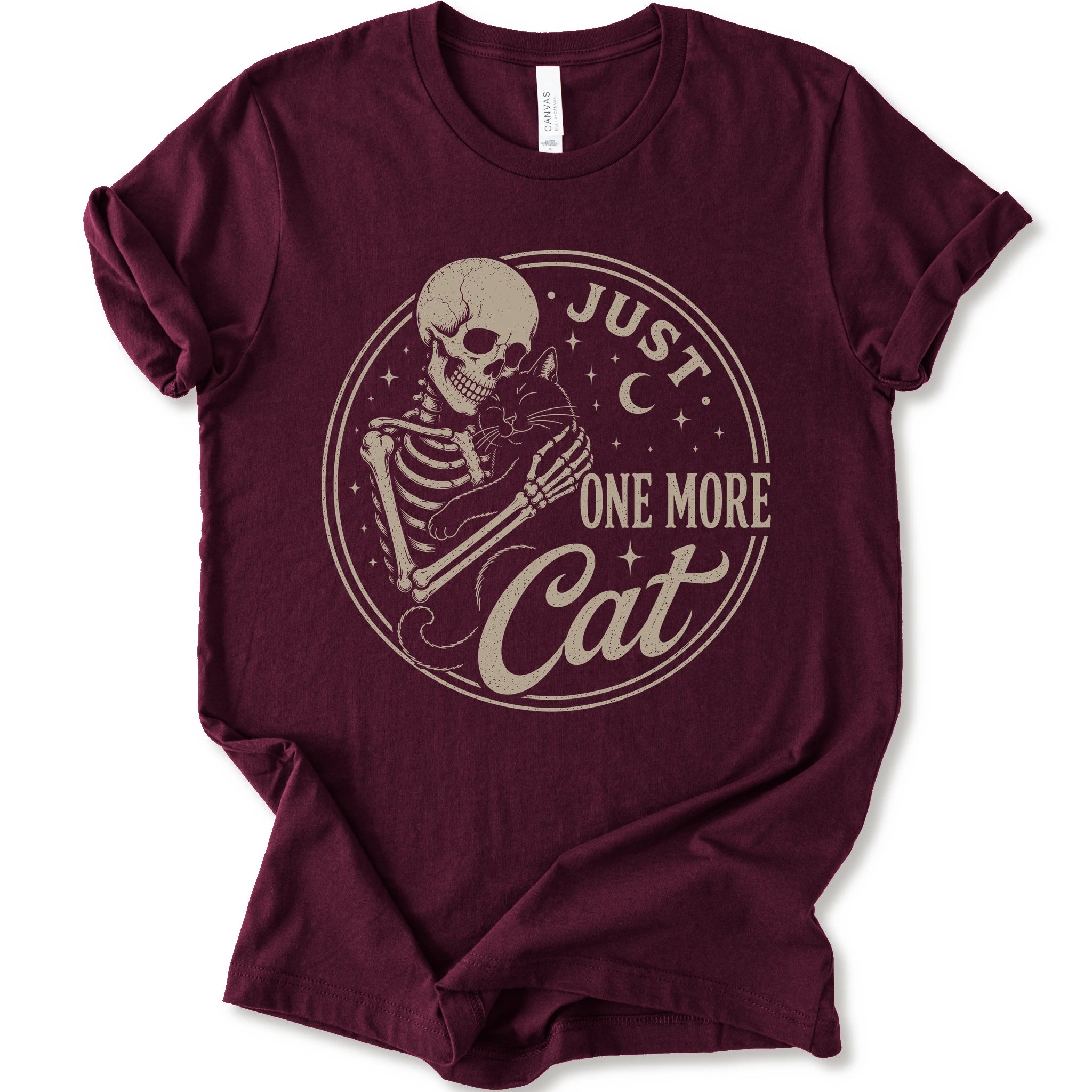 A maroon short-sleeve t-shirt with a vintage-style graphic of a skeleton holding a cat inside a circular frame with the text 'JUST ONE MORE Cat'.