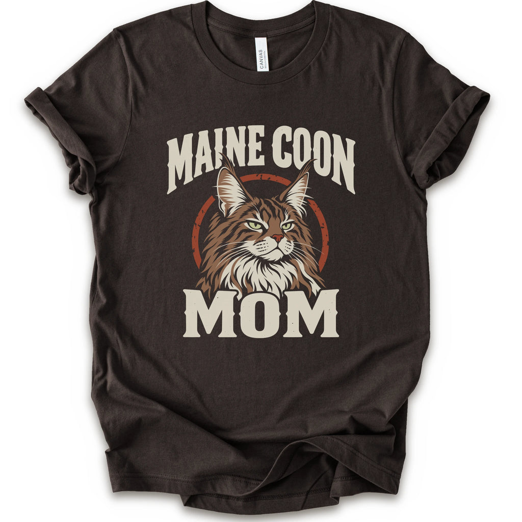 A brown short-sleeve crewneck t-shirt featuring a graphic print of a Maine Coon cat with the text 'MAINE COON MOM' in distressed cream lettering.