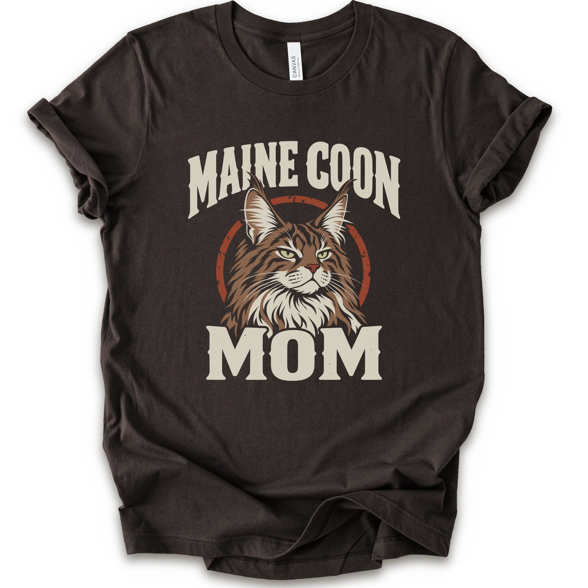 A brown short-sleeve crewneck t-shirt featuring a graphic print of a Maine Coon cat with the text 'MAINE COON MOM' in distressed cream lettering.