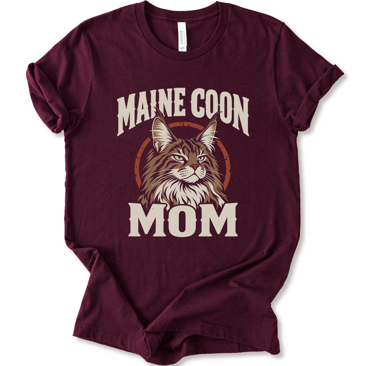 A maroon short-sleeve crewneck t-shirt featuring a graphic print of a Maine Coon cat with the text 'MAINE COON MOM' in distressed cream lettering.