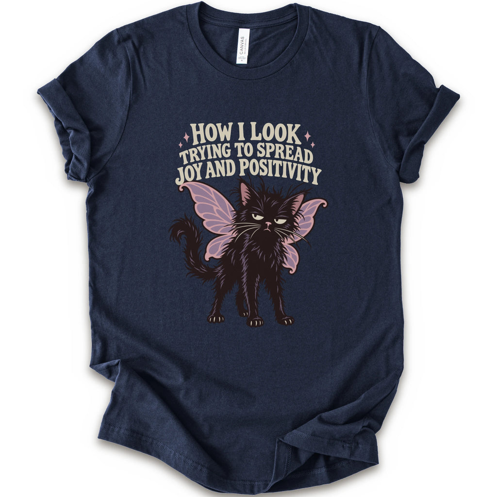 A navy short-sleeve crewneck t-shirt with a graphic of a black cat with pink-purple fairy wings and the text 'HOW I LOOK TRYING TO SPREAD JOY AND POSITIVITY' in a light beige font.