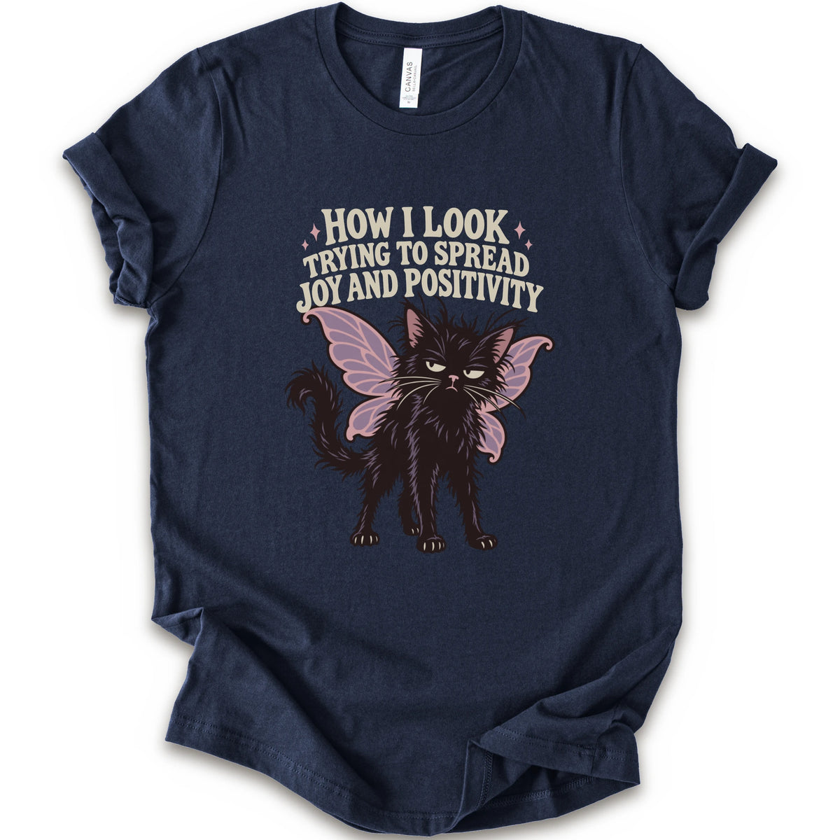 A navy short-sleeve crewneck t-shirt with a graphic of a black cat with pink-purple fairy wings and the text 'HOW I LOOK TRYING TO SPREAD JOY AND POSITIVITY' in a light beige font.