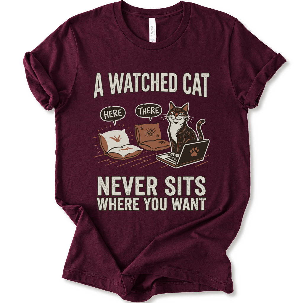A maroon short-sleeve crewneck t-shirt with a humorous graphic print of a cat on a laptop and couch, featuring the text 'A WATCHED CAT NEVER SITS WHERE YOU WANT' and speech bubbles reading 'HERE' and 'THERE'.