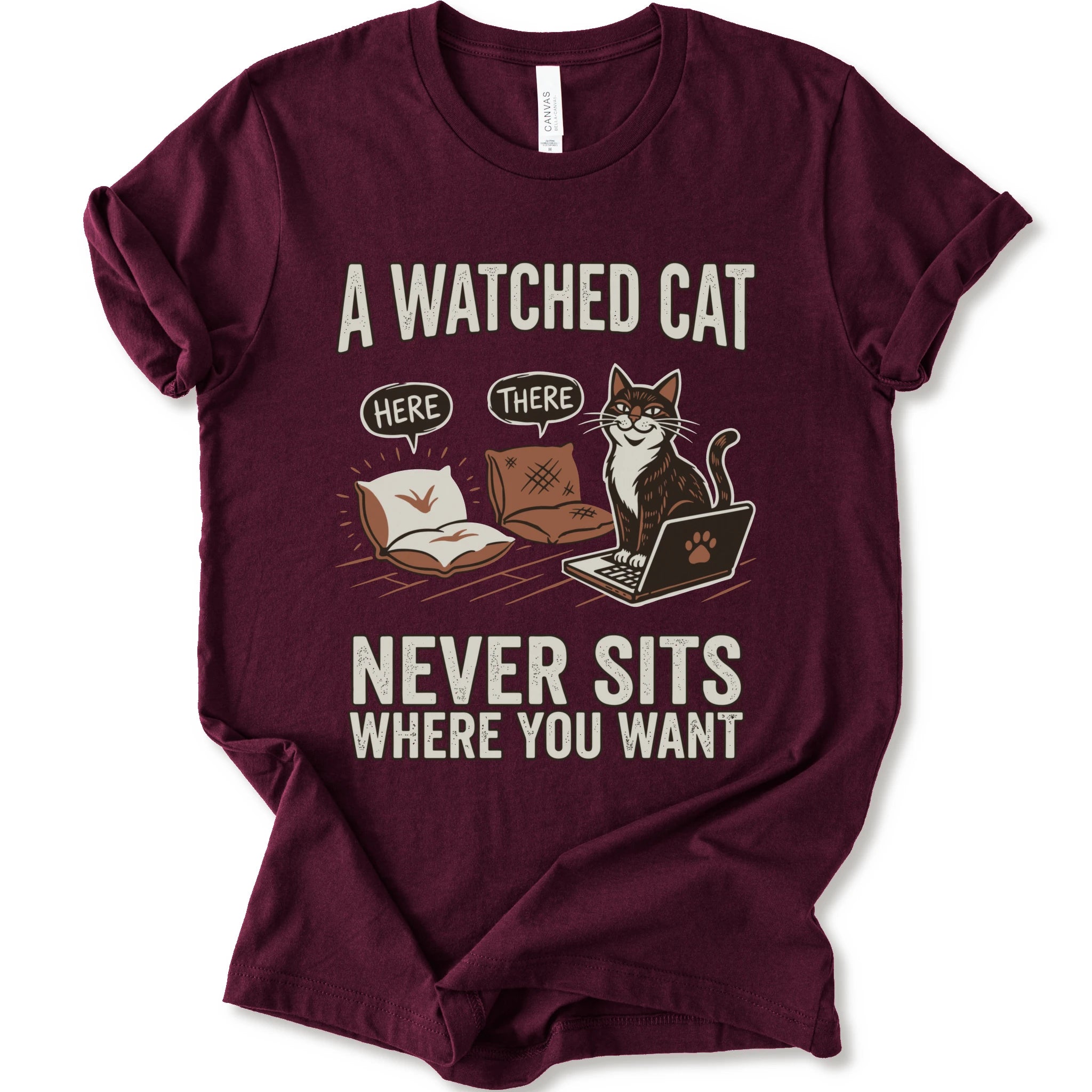 A maroon short-sleeve crewneck t-shirt with a humorous graphic print of a cat on a laptop and couch, featuring the text 'A WATCHED CAT NEVER SITS WHERE YOU WANT' and speech bubbles reading 'HERE' and 'THERE'.