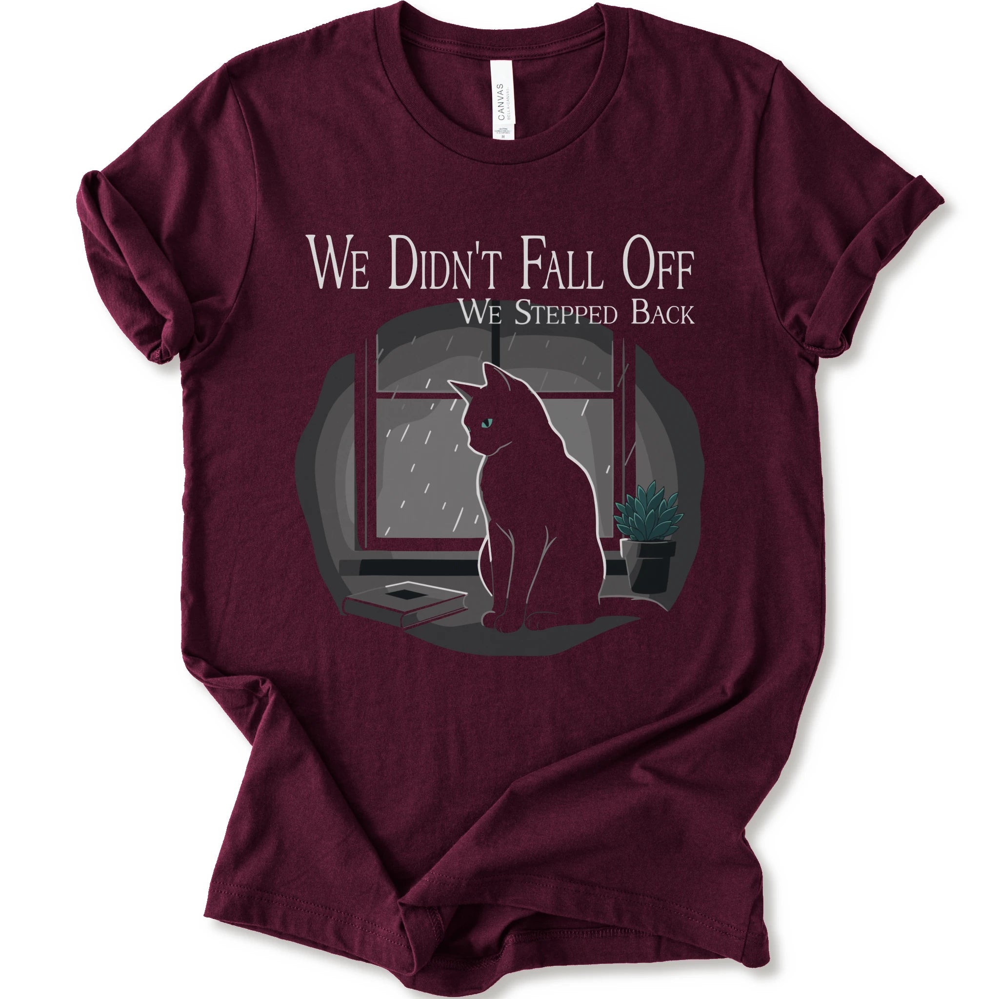 A maroon short-sleeve crewneck t-shirt with a graphic print of a black cat sitting in front of a window with the text 'WE DIDN'T FALL OFF WE STEPPED BACK'.