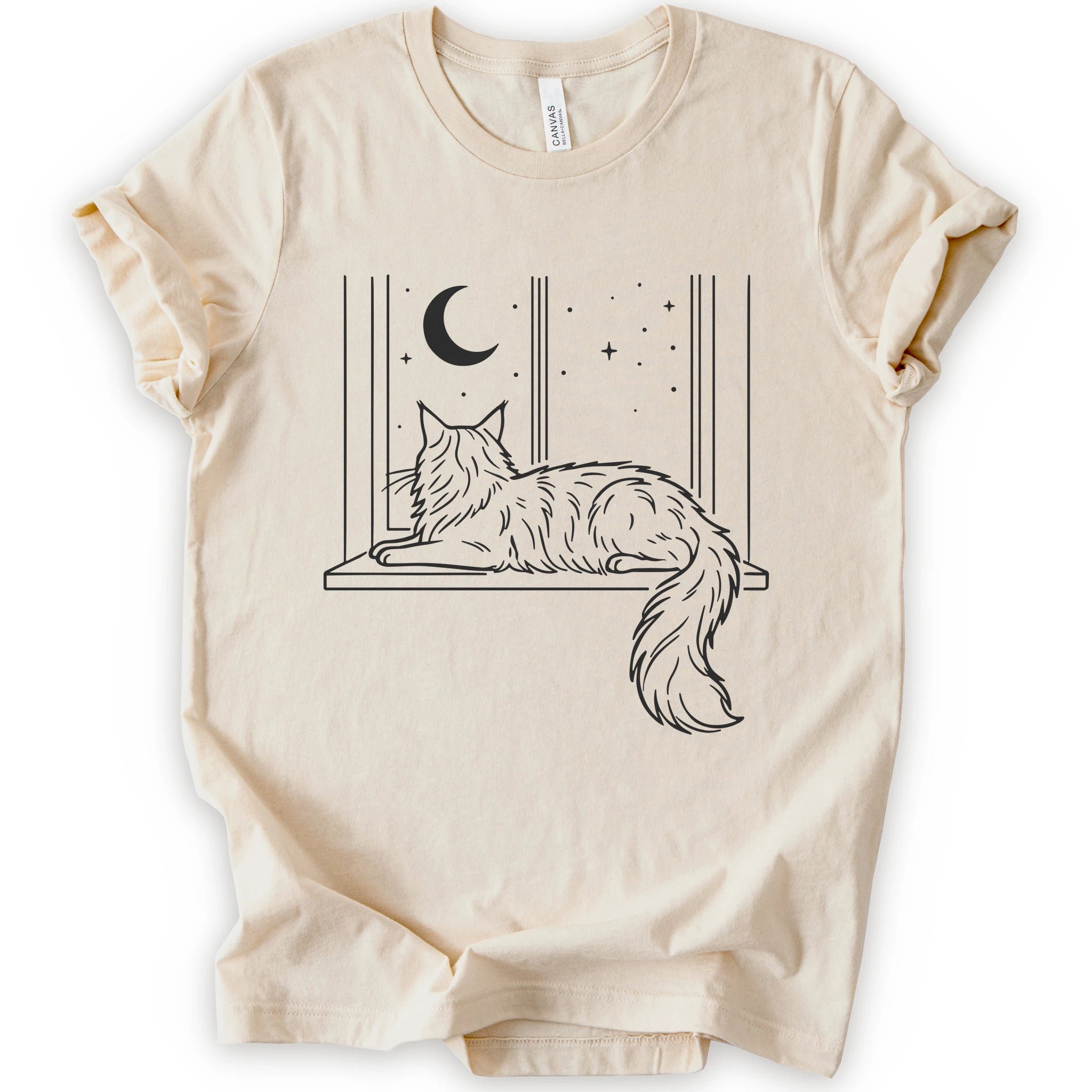 A natural short-sleeve crewneck t-shirt with a graphic print of a long-haired maine coon resting on a windowsill under a crescent moon and stars.