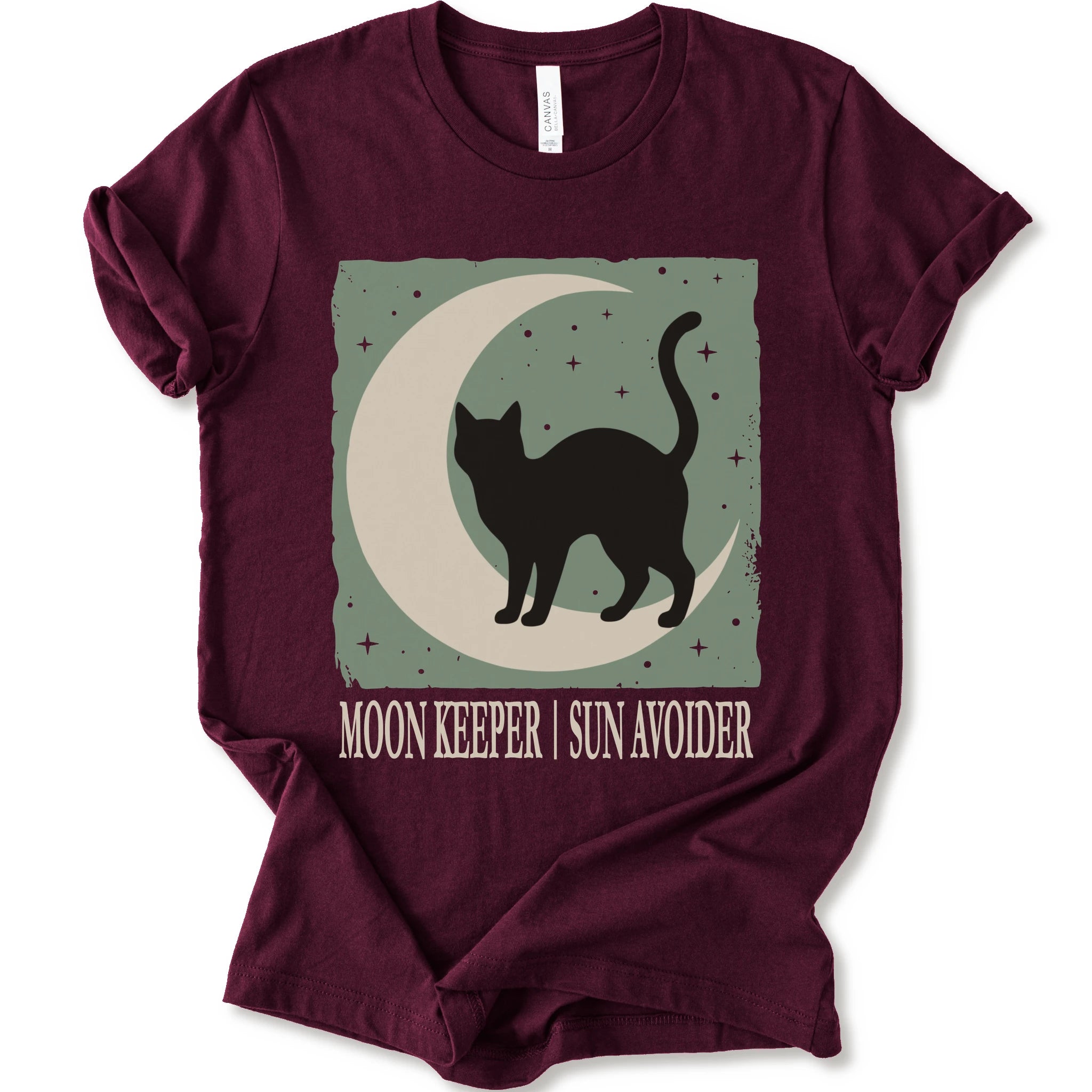 A maroon short-sleeve crewneck t-shirt with a graphic print of a black cat silhouette walking on a crescent moon against a sage green starry background, with the text 'MOON KEEPER | SUN AVOIDER' printed below.