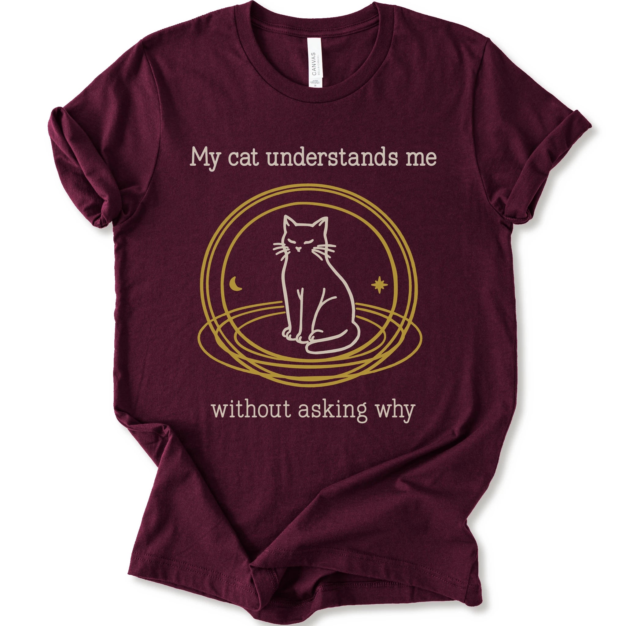 A maroon t-shirt with a graphic of a white cat sitting inside a gold circular frame, with the text 'My cat understands me without asking why' in a light beige font.