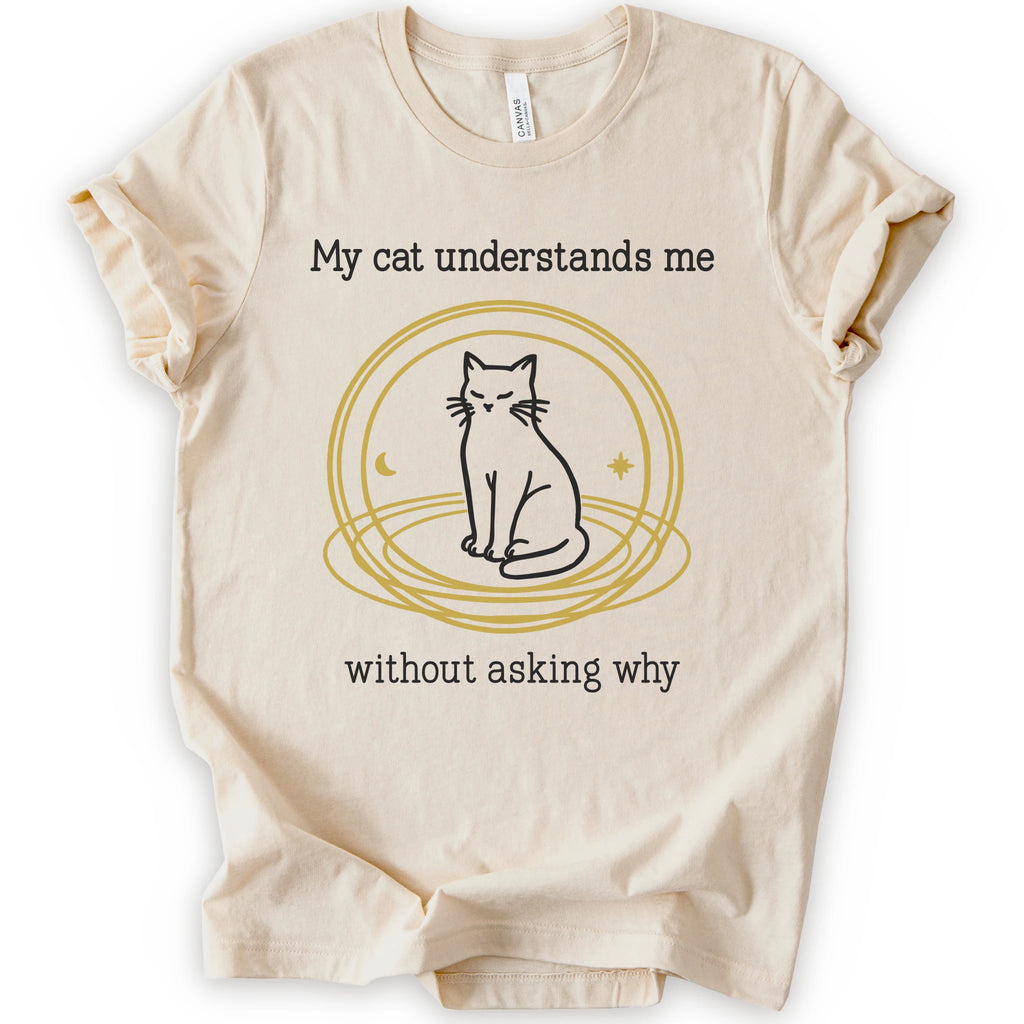 A natural t-shirt with a graphic of a black cat sitting inside a gold circular frame, with the text 'My cat understands me without asking why' in a black font.