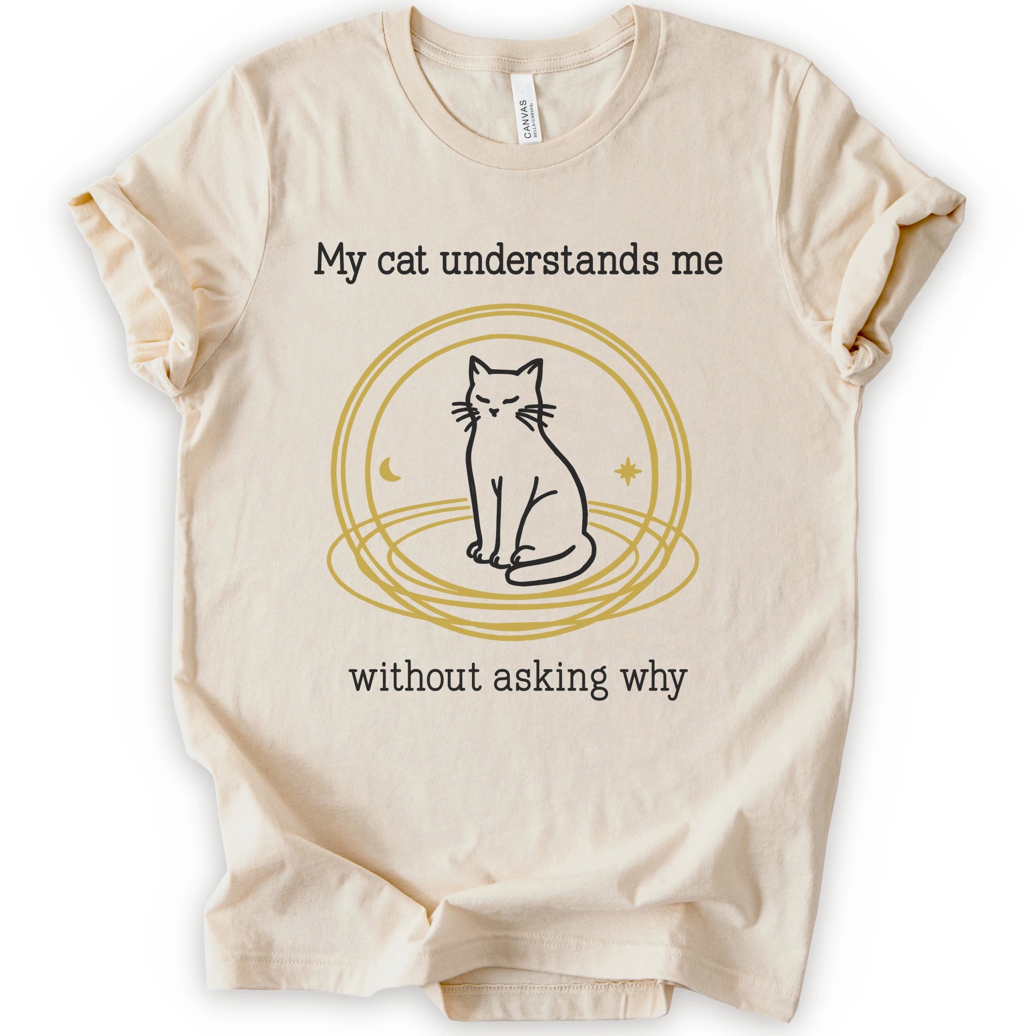 A natural t-shirt with a graphic of a black cat sitting inside a gold circular frame, with the text 'My cat understands me without asking why' in a black font.