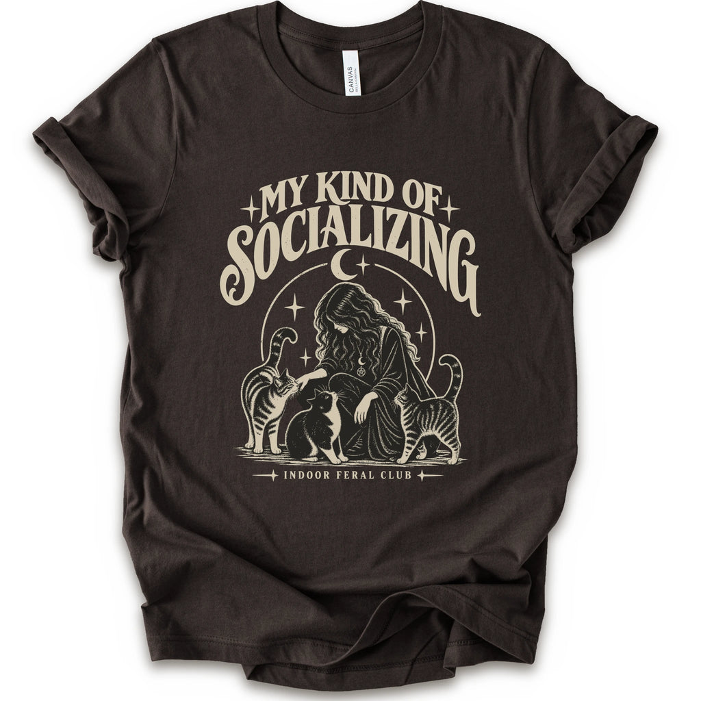 A brown short-sleeve t-shirt with a cream-colored graphic print on the front. The graphic features the text 'MY KIND OF SOCIALIZING' in a stylized font above an illustration of a woman surrounded by cats under a crescent moon, with 'INDOOR FERAL CLUB' printed below.