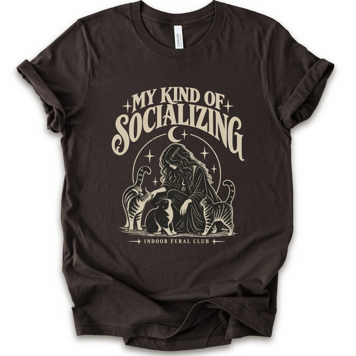 A brown short-sleeve t-shirt with a cream-colored graphic print on the front. The graphic features the text 'MY KIND OF SOCIALIZING' in a stylized font above an illustration of a woman surrounded by cats under a crescent moon, with 'INDOOR FERAL CLUB' printed below.