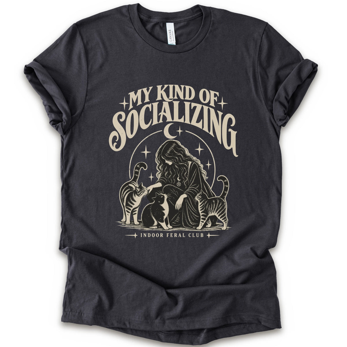 A dark grey short-sleeve t-shirt with a cream-colored graphic print on the front. The graphic features the text 'MY KIND OF SOCIALIZING' in a stylized font above an illustration of a woman surrounded by cats under a crescent moon, with 'INDOOR FERAL CLUB' printed below.