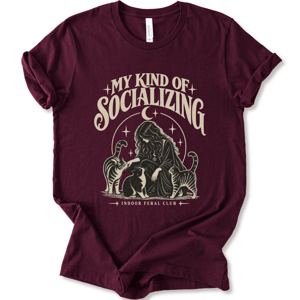 A maroon short-sleeve t-shirt with a cream-colored graphic print on the front. The graphic features the text 'MY KIND OF SOCIALIZING' in a stylized font above an illustration of a woman surrounded by cats under a crescent moon, with 'INDOOR FERAL CLUB' printed below.