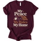 A maroon short-sleeve crewneck t-shirt with a retro-style graphic print that reads 'My Peace My Rules My Home' in cream and brown text, featuring an illustration of a black cat under a brown blanket next to a small mug.