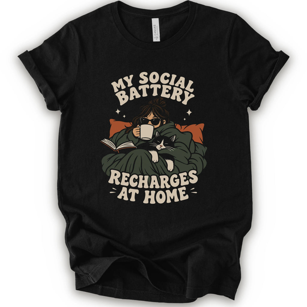 A black short-sleeve graphic t-shirt features a cozy illustration of a person wrapped in a green blanket reading a book with a black cat, surrounded by the text 'MY SOCIAL BATTERY RECHARGES AT HOME' in a beige retro font.